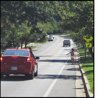 Both photos show the same crosswalk at a narrow two-lane residential street.  About 150 feet before the crosswalk we see a post with two signs on it - the top sign shows a drawing of a person walking, below that is a sign saying deaf/blind 'pedestrians.' Standing on the right side of the street is a woman wearing white shorts and a red-dand-white-checkered blouse, standing with one foot in front of the other and holding a white cane with the tip on the ground.  A red car has stopped just before reaching the sidewalk.